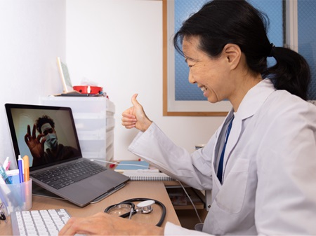 Smiling provider sitting in front of laptop gestures a thumbs up to the masked patient on her screen during a virtual visit