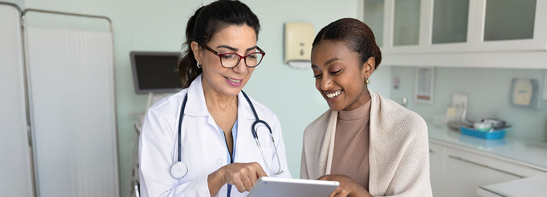 A female doctor, sitting across a table from a patient, places her hands on top of her patient's hands, offering comfort.